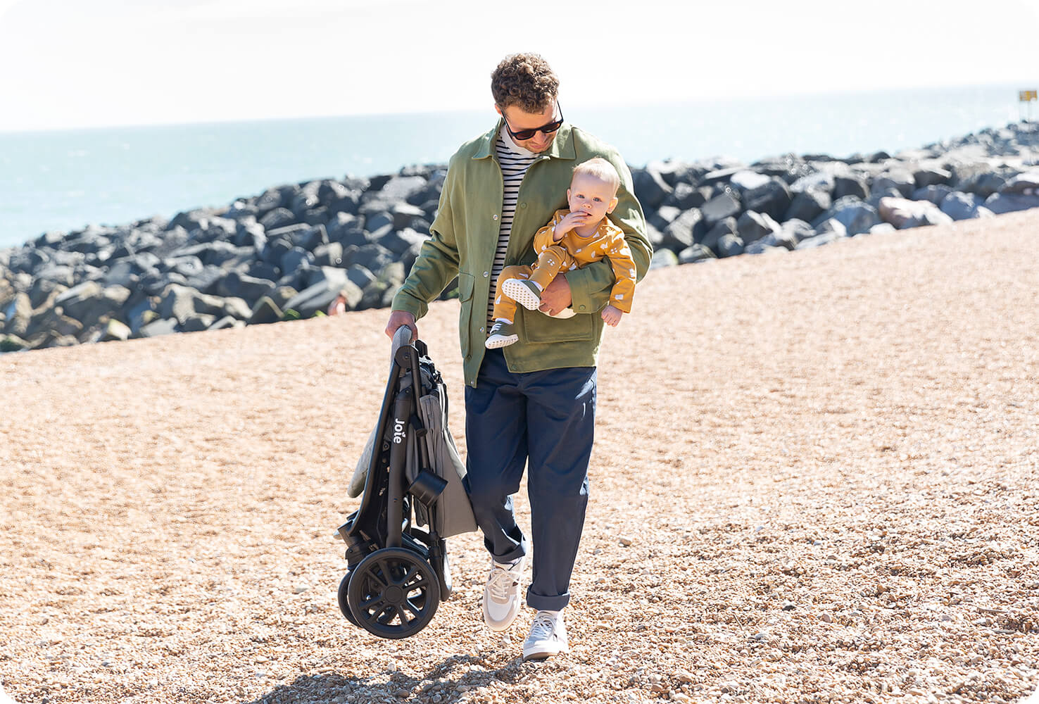  Père marchant sur une plage, portant un bébé à un bras et une poussette litetrax 4 dlx repliée dans l’autre main.