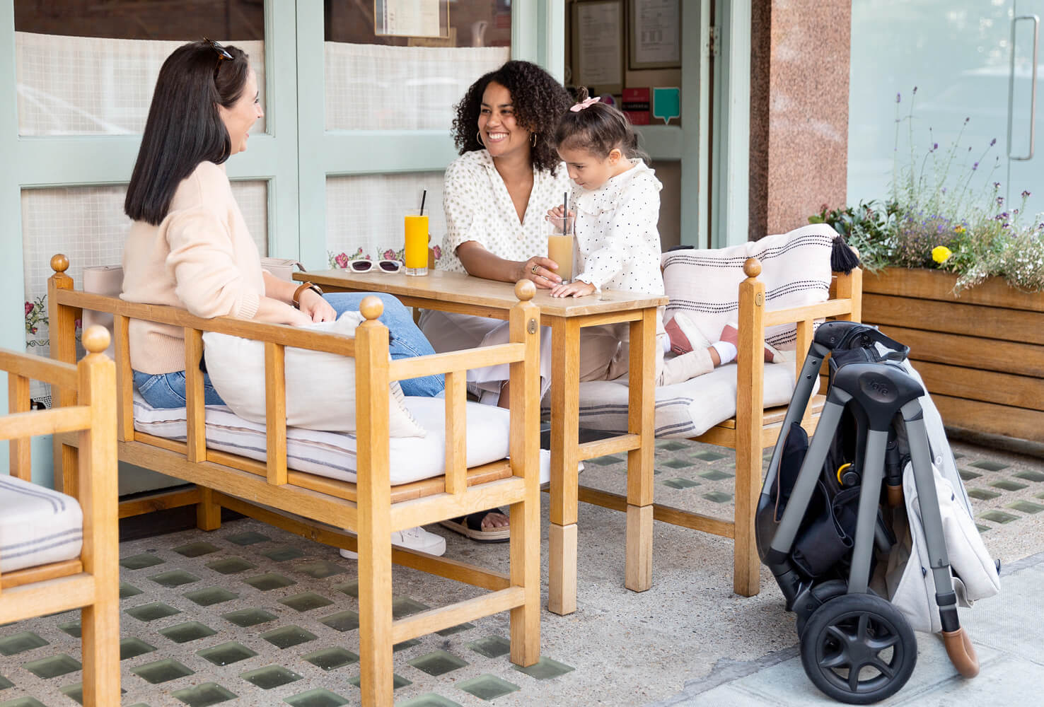  Joie finiti pram in light gray folded leaning against table as toddler sits on moms lap while talking to another woman.