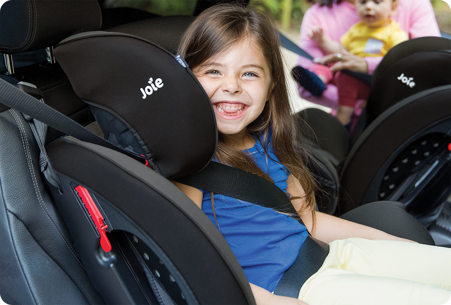 A young girl sitting in a black Joie Stages FX car seat in booster mode, smiling at the camera with a mum and baby in the background on the other side of the car