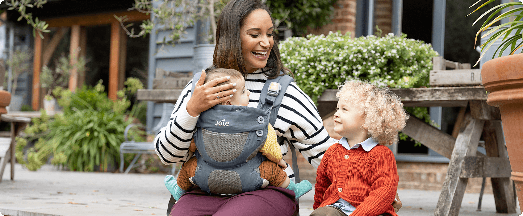 A mum and toddler sitting on a patio. Mum is wearing a baby in a blue Joie Savvy baby carrier.