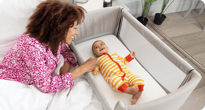 Mum lying in bed smiling at baby who is lying in the Joie Roomie Go bedside bassinet